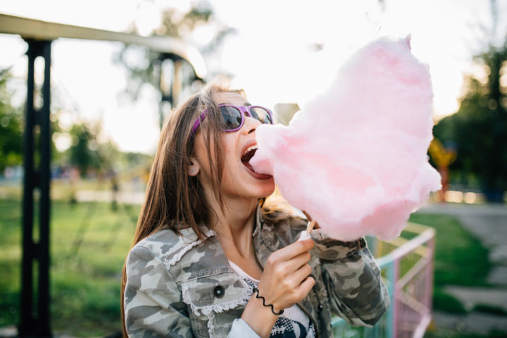 Outdoor photo of a young trendy woman in sunglasses eating a cotton candy, spending tie with pleasure. Dressed in fashionable jacket.